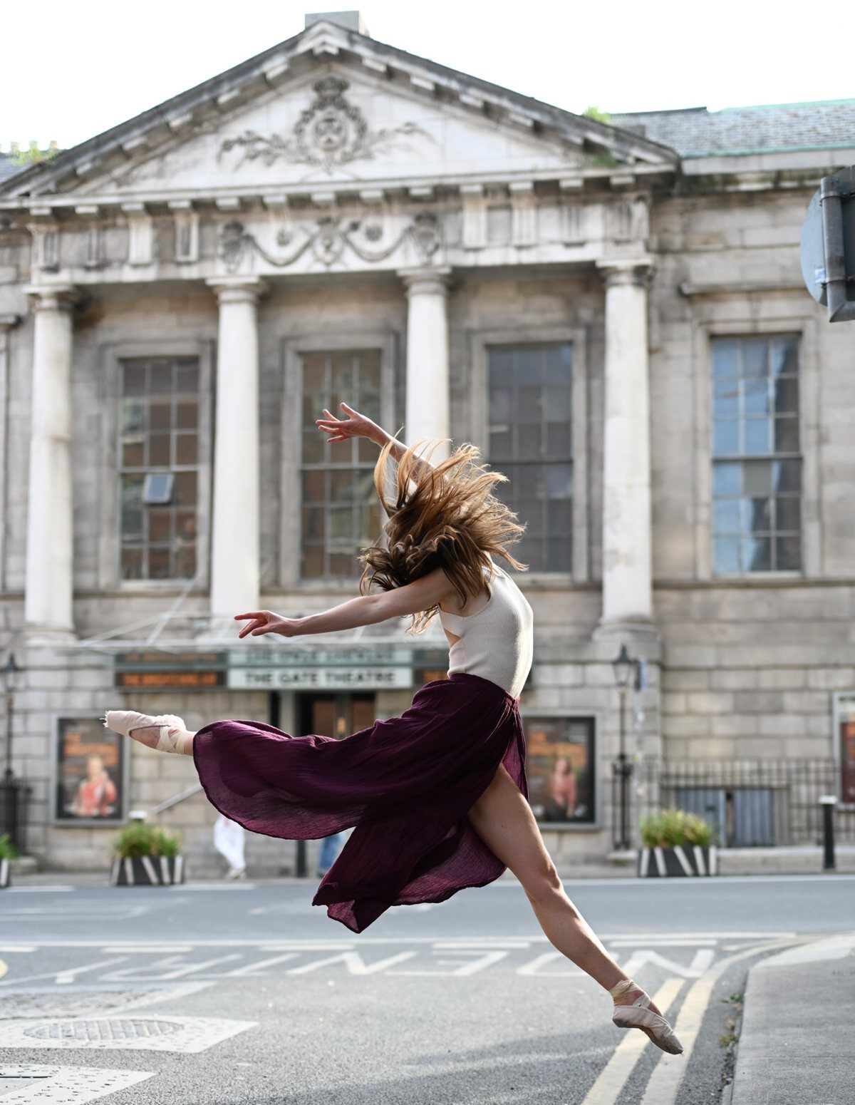 Dance artist Laura Sarah Dowdall leaps across the frame outside The Gate Theatre in Dublin, her legs and arms extended and her long hair lifted by the movement. She wears ballet shoes, a white sleeveless top and a flowing plum skirt. The theatre’s columned façade and “The Gate Theatre” sign are visible behind her.
